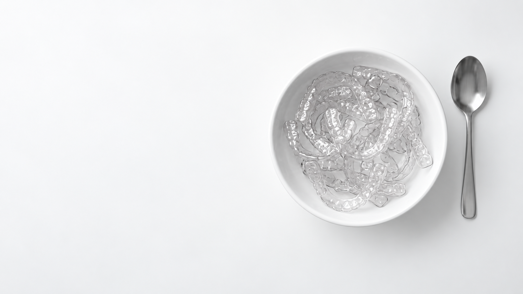 A white bowl filled with clear retainers beside a stainless steel spoon